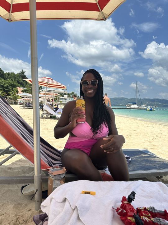 Woman with a cocktail under a striped umbrella, boats on turquoise water at Doctor's Cave, Montego Bay